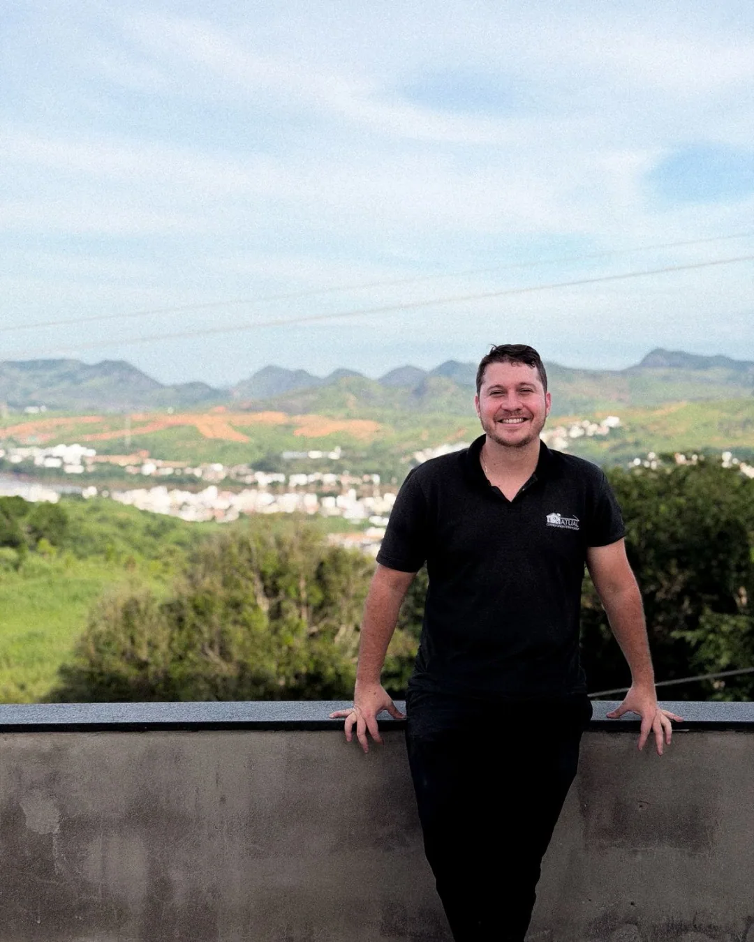 Homem de camisa polo com a marca Atual, sorrindo em um mirante com a cidade e as montanhas ao fundo.
